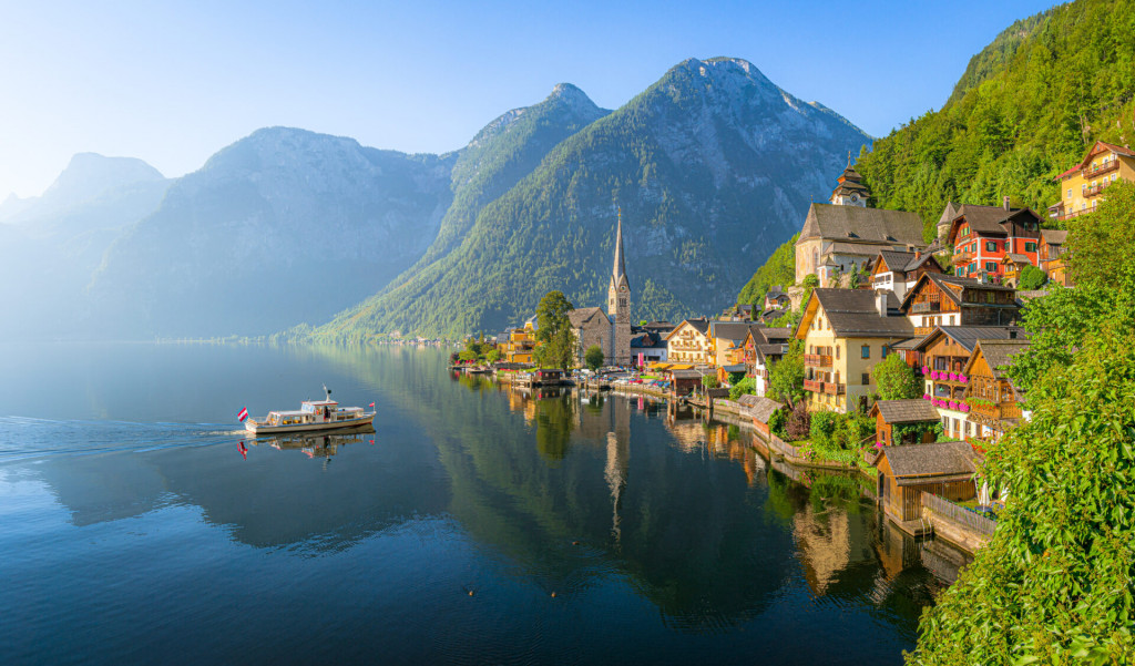 Classic panoramic view of idyllic Hallstatt lake in the Alps with famous old town with tourist ship in scenic golden morning light on a beautiful sunny day at sunrise in summer, Salzkammergut region, Austria